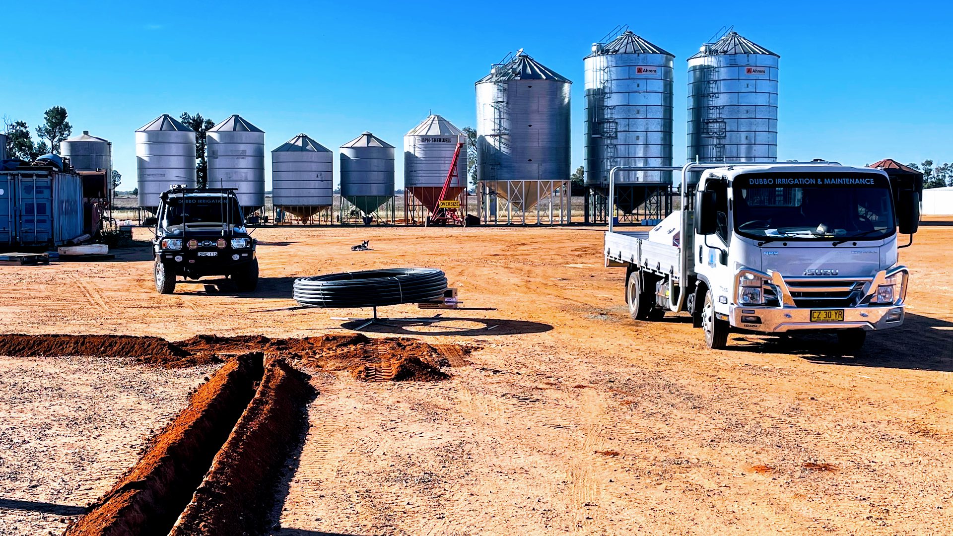 Grain silos and trucks behind a fresh trench in red soil