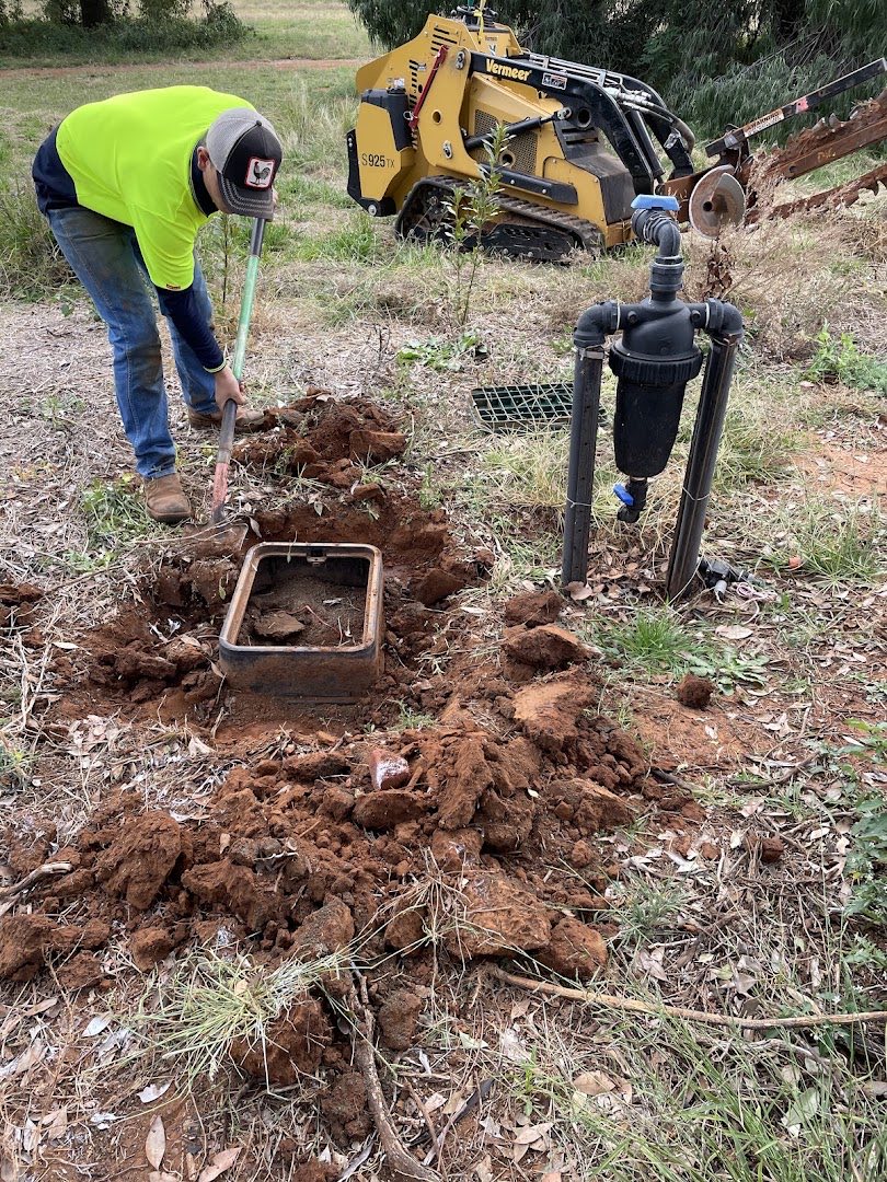 A worker in a hi-vis shirt digging a valve box into red soil, a mini loader behind him