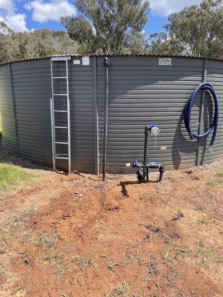 A tall corrugated steel water tank with a ladder, standing on freshly turned ground