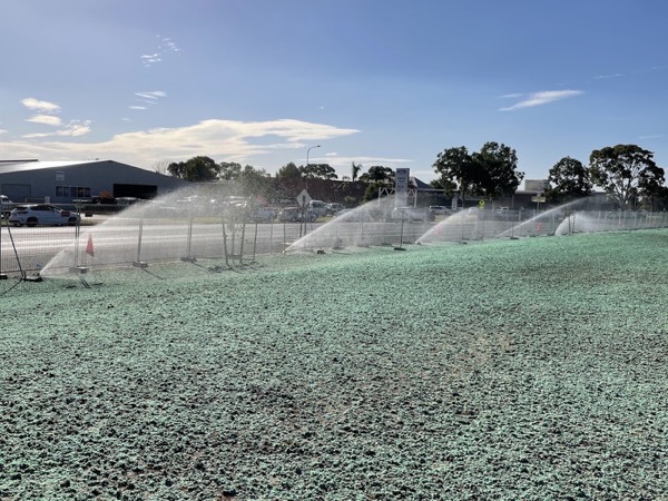 Sprinklers throwing water across a large green field with sheds behind