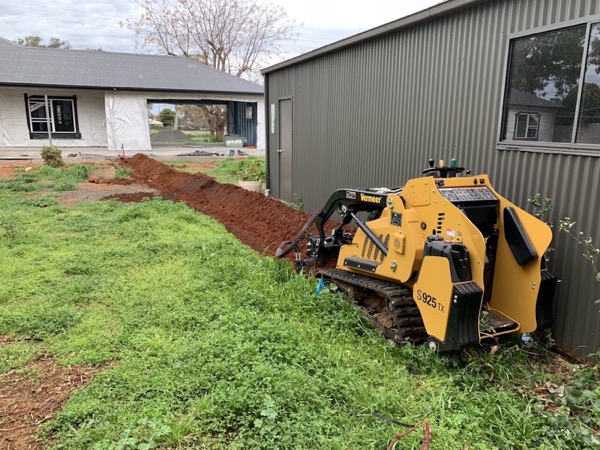 A tracked mini loader cutting a trench along the edge of a green lawn