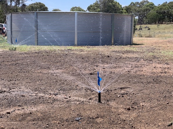 A single sprinkler running over freshly graded bare ground with a steel tank behind