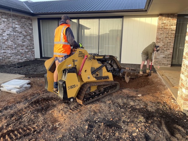 A worker operating a tracked mini loader beside a house while another shovels soil