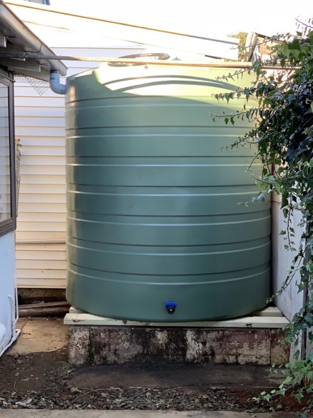 A green poly water tank on a concrete base beside a weatherboard wall