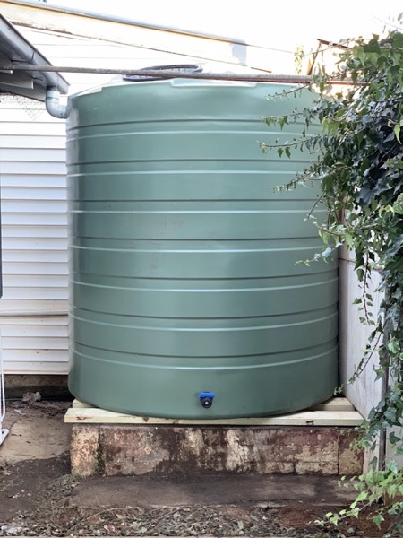 A green poly water tank on a raised base beside a house
