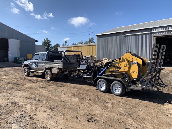 A ute and trailer with a tracked mini loader parked beside farm sheds