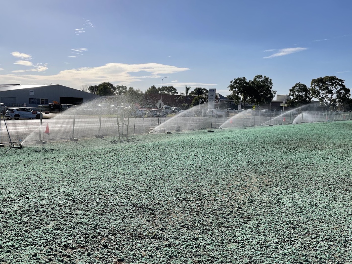 Sprinklers throwing water across a large green field with sheds behind